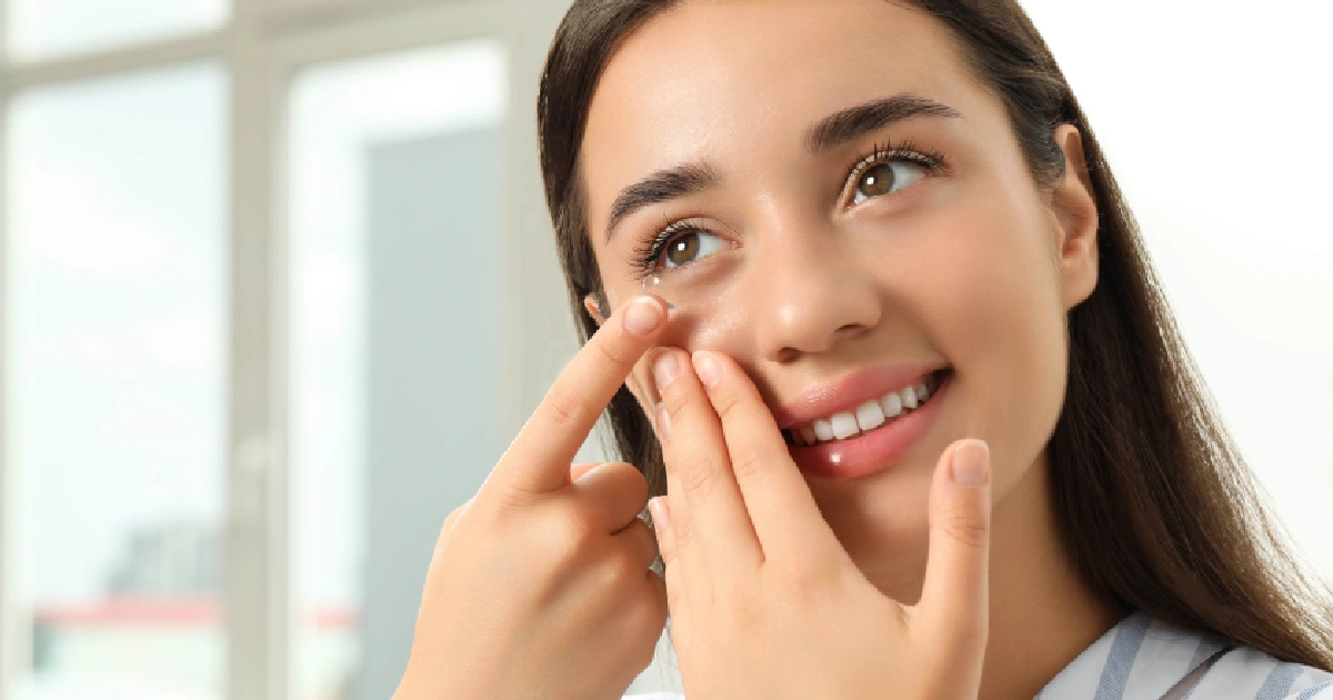 Close-up of a smiling woman inserting a custom contact lens in Glenview, IL, with hands steadying her eyelid and a bright window behind her.